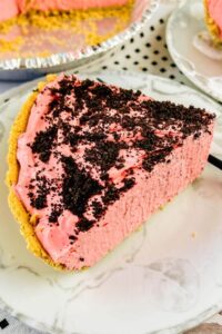 A slice of raspberry cream pie with a graham cracker crust and chocolate cookie crumbs on top, served on a white plate.