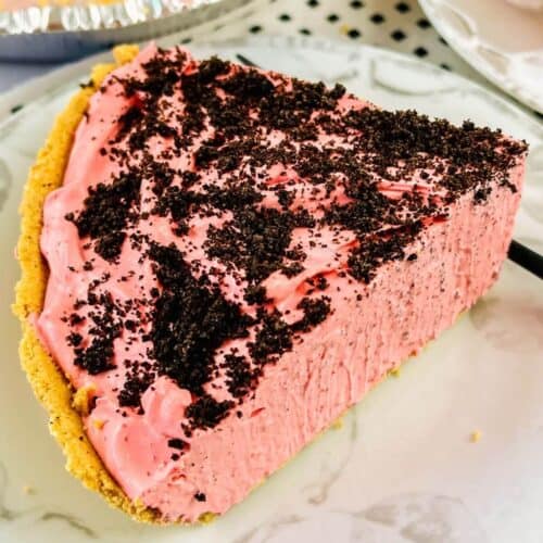 A slice of raspberry cream pie with a graham cracker crust and chocolate cookie crumbs on top, served on a white plate.