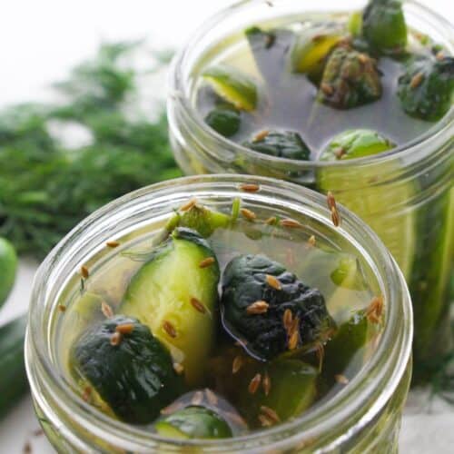 Two glass jars filled with sliced cucumbers in brine, garnished with dill, seeds, and garlic, create the perfect Garlic Dill Pickles, set against a white background.