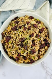 A bowl of wild rice salad with dried cranberries, chopped vegetables, and herbs is served as a vibrant rice dressing on a white surface, accented by a striped cloth in the background.