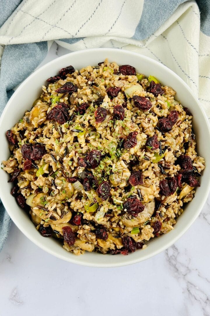 A bowl of wild rice salad with dried cranberries, chopped vegetables, and herbs is served as a vibrant rice dressing on a white surface, accented by a striped cloth in the background.