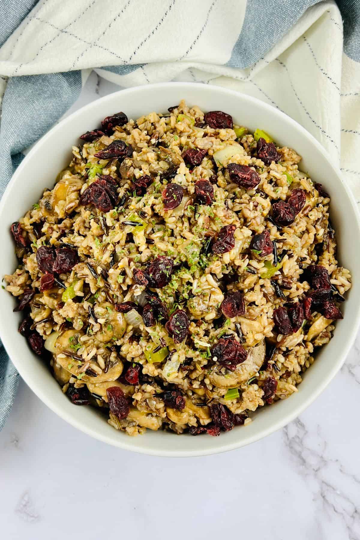 A bowl of wild rice salad with dried cranberries, chopped vegetables, and herbs is served as a vibrant rice dressing on a white surface, accented by a striped cloth in the background.