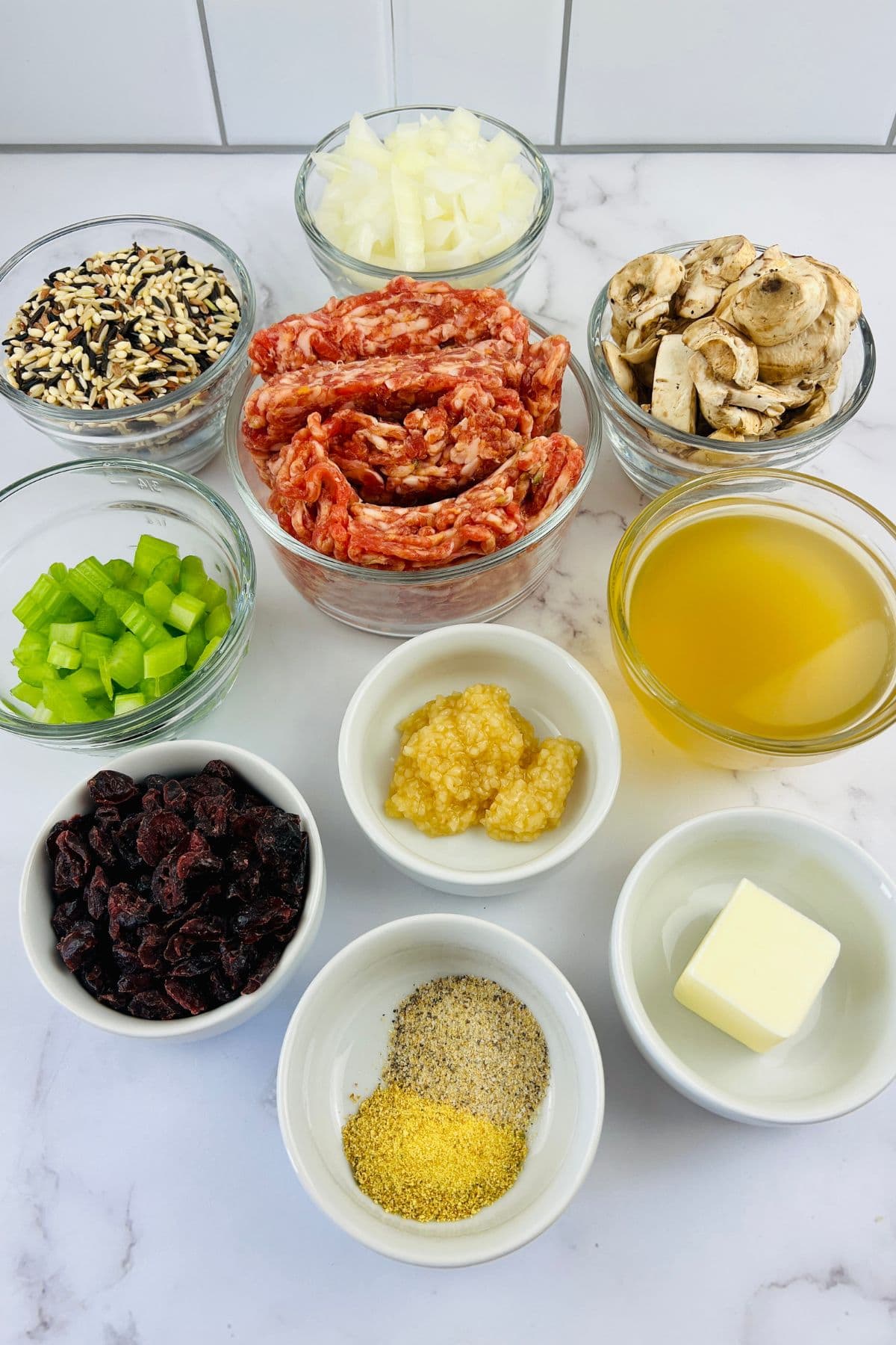 Nine bowls contain ingredients  raw sausage, diced onions, sliced mushrooms, wild rice blend, chopped celery, chicken broth, minced garlic, dried cranberries, seasonings, and a pat of butter.