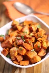 A white bowl filled with Brown Sugar Sweet Potatoes, garnished with chopped herbs, sits on a wooden surface with a spoon and orange napkin in the background.