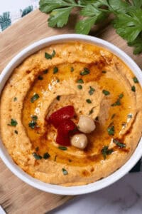 A bowl of roasted red pepper hummus garnished with chickpeas, olive oil, and parsley sits invitingly on a wooden board.