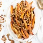 Oval dish of Roasted Spicy Sweet Potatoes wedges topped with chopped pecans, with extra pecans scattered on a white marble surface nearby.