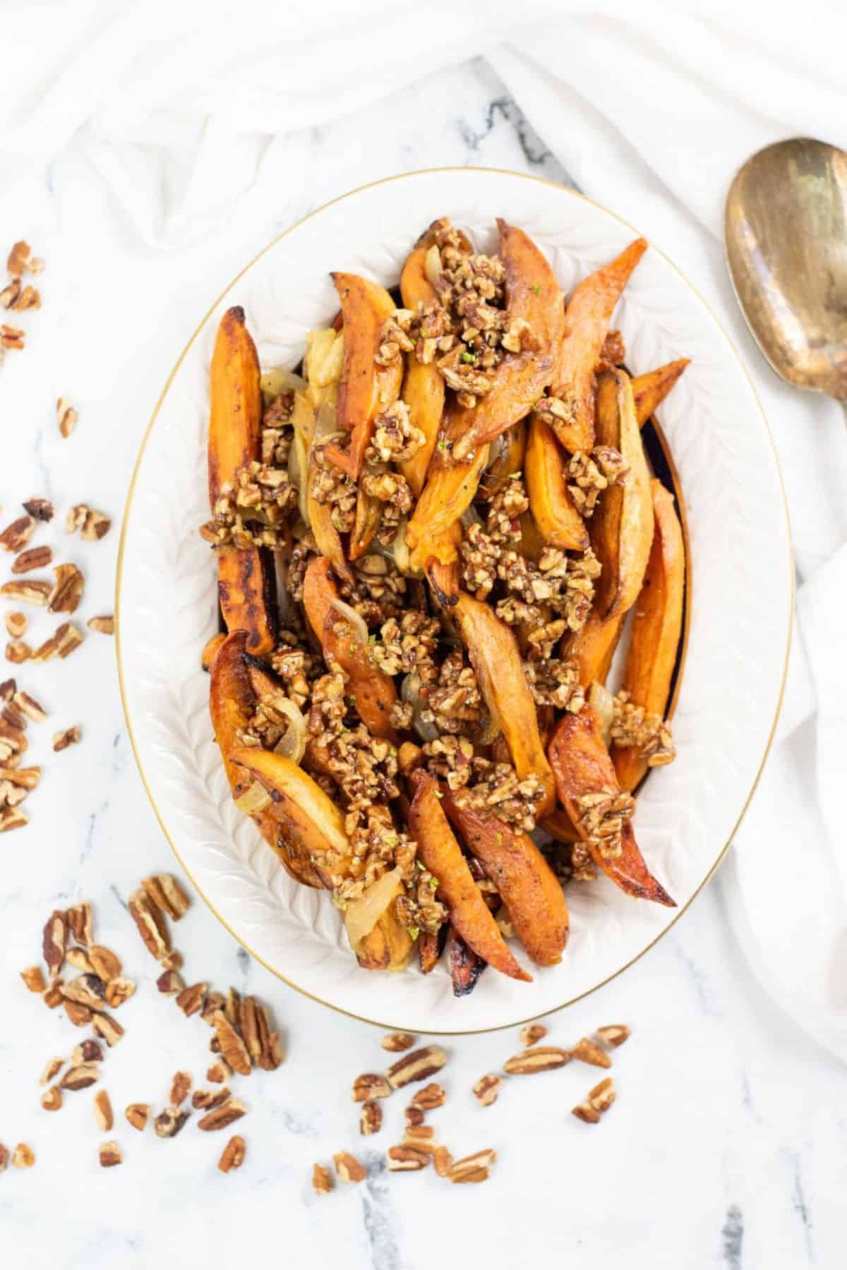 Oval dish of Roasted Spicy Sweet Potatoes wedges topped with chopped pecans, with extra pecans scattered on a white marble surface nearby.