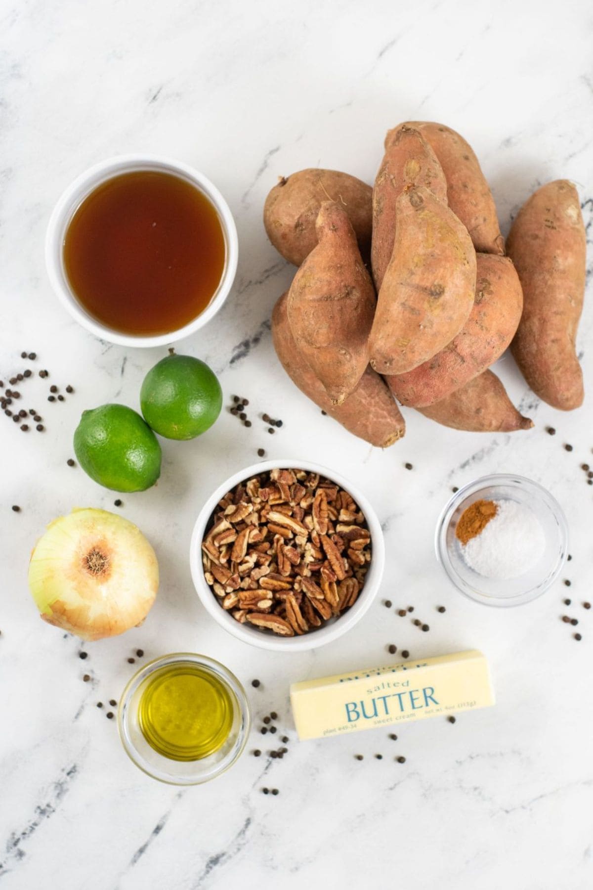 Assorted ingredients for Roasted Spicy Sweet Potatoes on a marble surface, including sweet potatoes, lime, pecans, onion, maple syrup, olive oil, butter, salt, black pepper, and ground cinnamon.