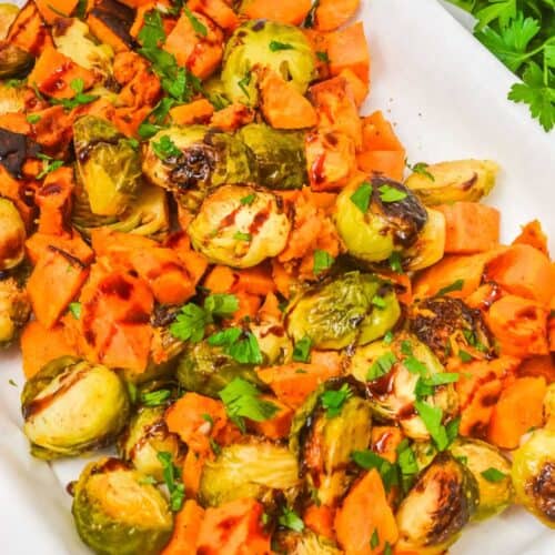Roasted Brussels sprouts and sweet potatoes, classic roasted vegetables, are garnished with chopped parsley and served on a parchment-lined tray.