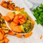 A serving spoon holds roasted Brussels sprouts and sweet potato cubes above a baking sheet lined with parchment paper. Fresh parsley is visible in the background, highlighting this colorful medley of roasted vegetables.