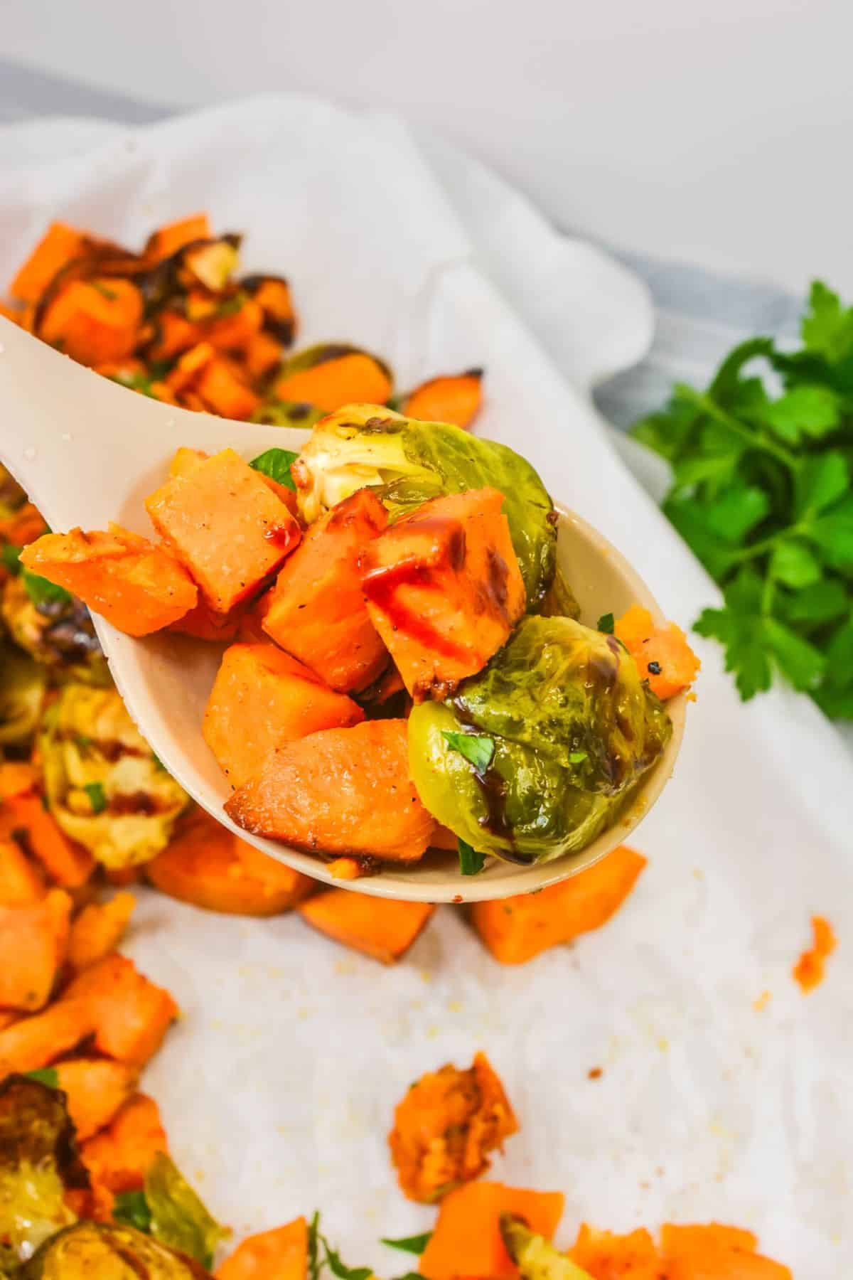 A serving spoon holds roasted Brussels sprouts and sweet potato cubes above a baking sheet lined with parchment paper. Fresh parsley is visible in the background, highlighting this colorful medley of roasted vegetables.
