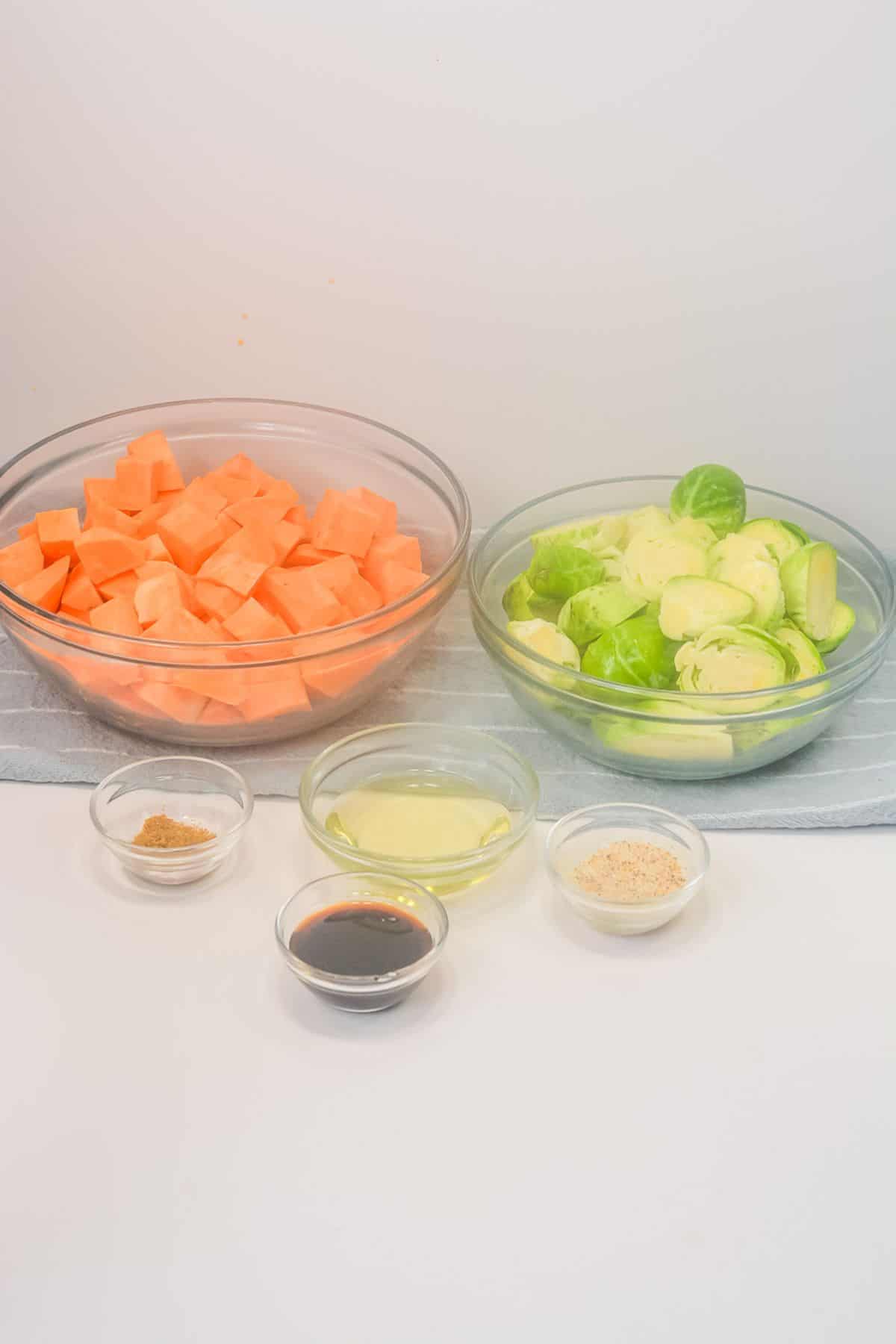 Two glass bowls with cubed vegetables, plus four small bowls containing oil, soy sauce, breadcrumbs, and seasoning, arranged on a white surface.