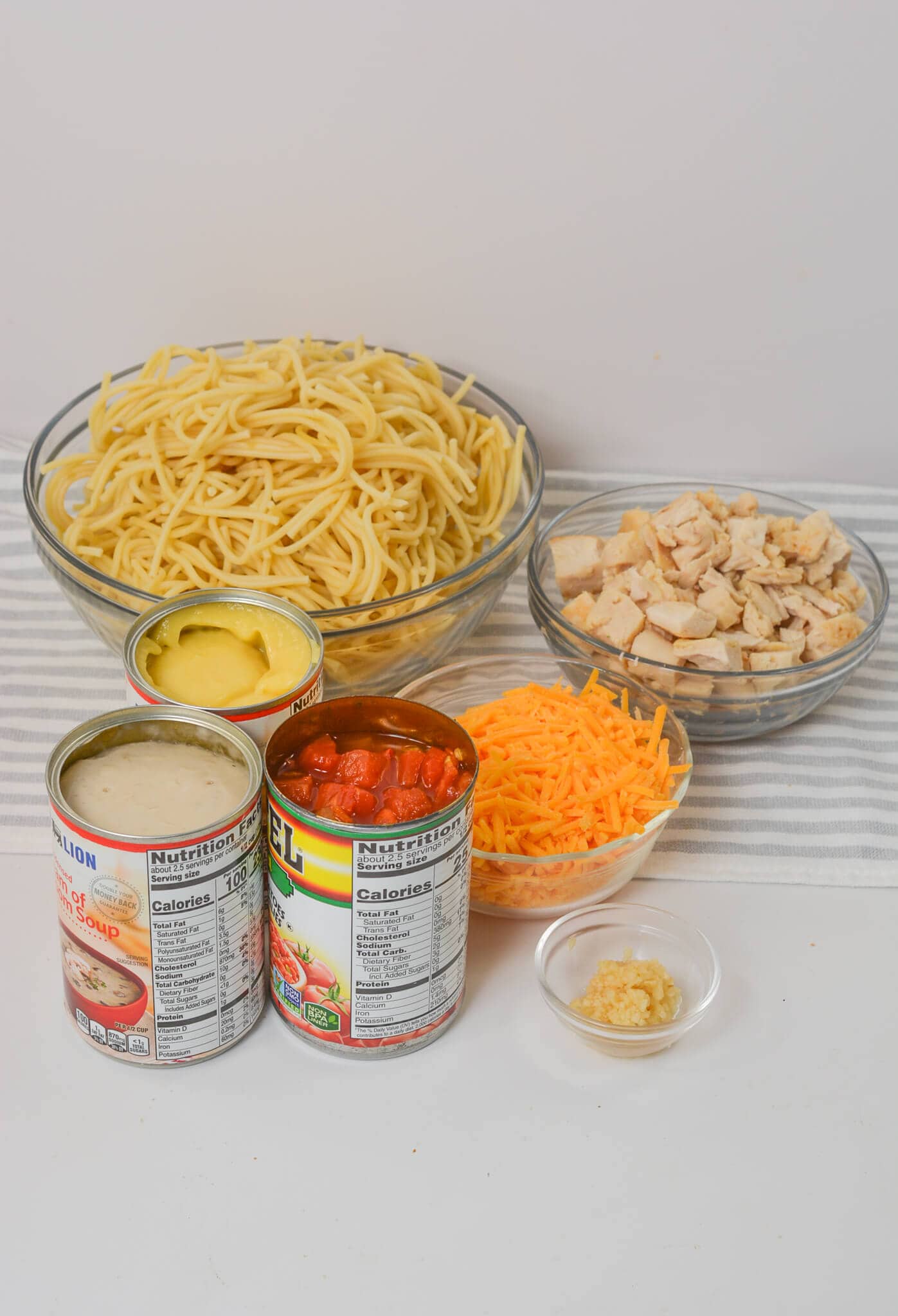 Ingredients on a table: bowls of spaghetti, cubed chicken, shredded cheese, and minced garlic; canned cream of chicken soup and a can of Rotel for that perfect kick.