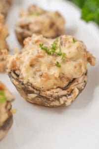 Cream cheese filled mushrooms are delicately topped with a creamy filling and garnished with chopped parsley, all elegantly arranged on a white plate.