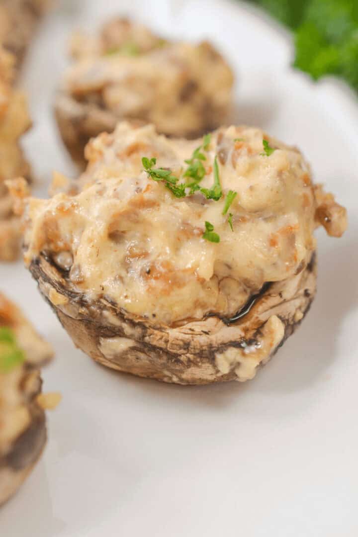 Cream cheese filled mushrooms are delicately topped with a creamy filling and garnished with chopped parsley, all elegantly arranged on a white plate.