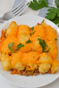 A plate of Sloppy Joe Tater Tot Casserole topped with melted cheddar cheese and garnished with chopped parsley. A fork and sprigs of parsley linger in the background, completing the inviting scene.