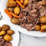 A white dish with Slow Cooker Beef Chuck Roast, baby potatoes, and carrots, next to a white plate holding a serving of beef and potatoes on a marble surface.