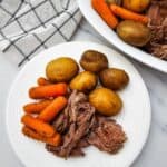 A white plate with Slow Cooker Beef Chuck Roast, cooked baby potatoes, and carrots, sits next to a serving dish with more of the hearty meal and a checkered napkin.
