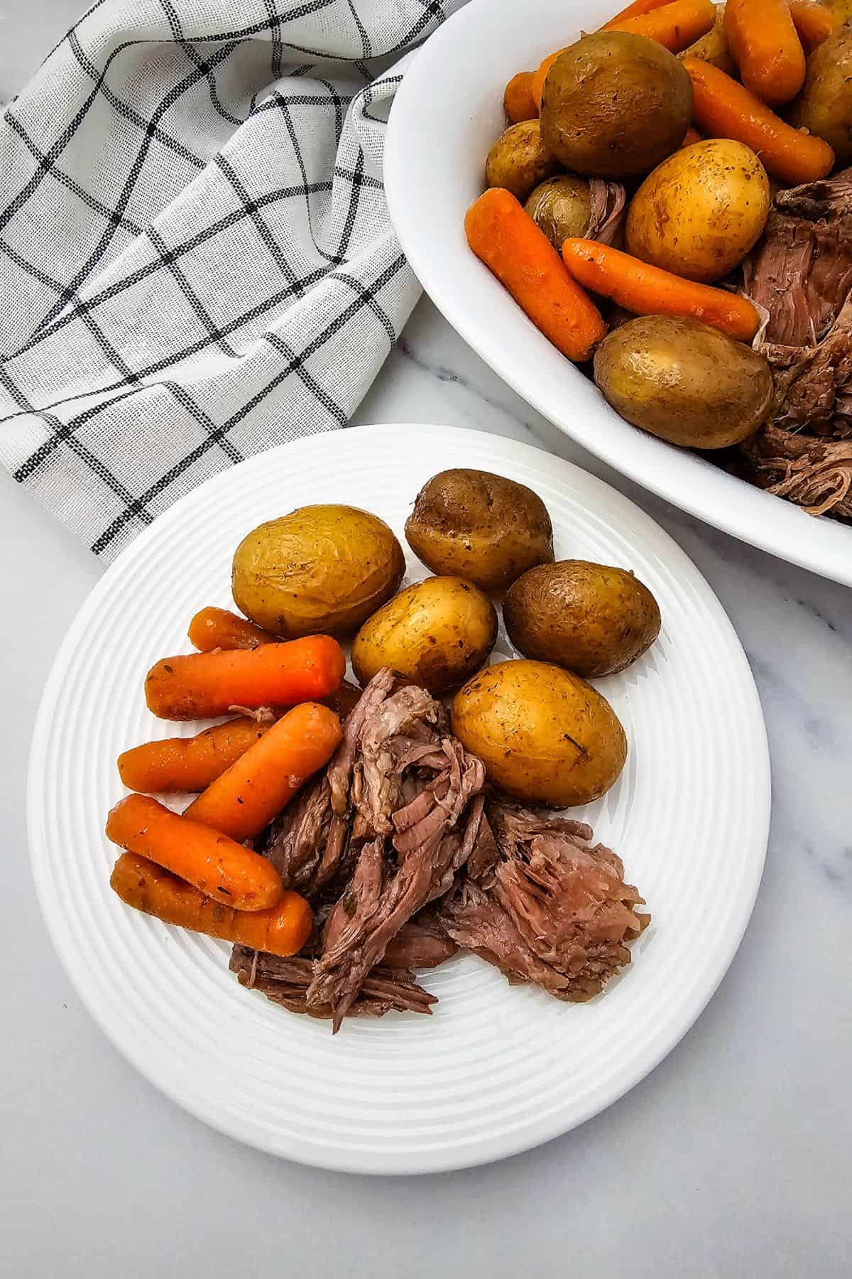 A white plate with Slow Cooker Beef Chuck Roast, cooked baby potatoes, and carrots, sits next to a serving dish with more of the hearty meal and a checkered napkin.