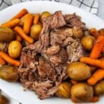 A white plate with shredded Slow Cooker Beef Chuck Roast, whole baby potatoes, and cooked carrots set on a marble surface next to a checkered cloth.