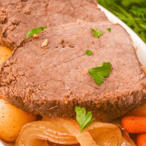 Plate of sliced slow cooker beef sirloin tip roast with potatoes, carrots, and onions, garnished with parsley.