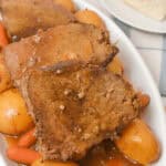 A plate of slow cooker beef sirloin tip roast slices is served with tender potatoes and carrots, all topped with a rich sauce. A slice of bread rests on a separate plate in the background, completing this hearty meal.