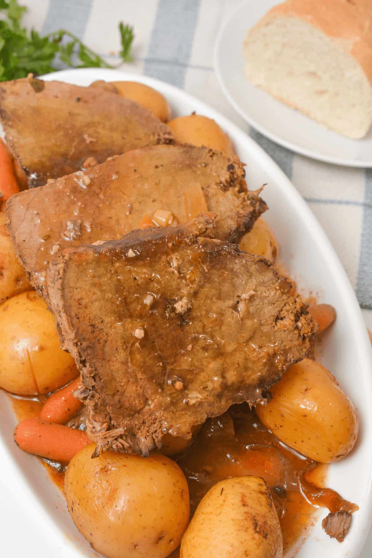 A plate of slow cooker beef sirloin tip roast slices is served with tender potatoes and carrots, all topped with a rich sauce. A slice of bread rests on a separate plate in the background, completing this hearty meal.