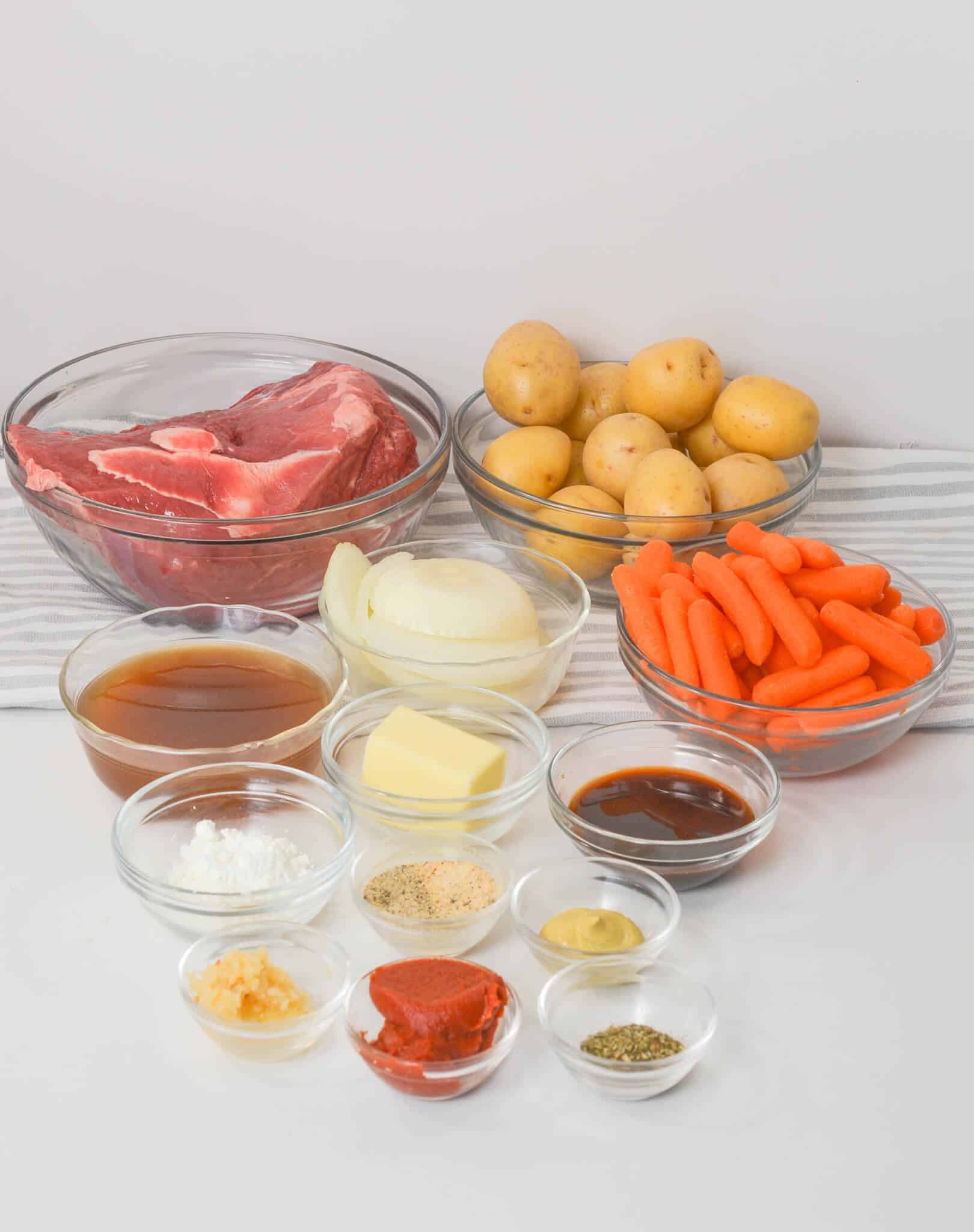 Ingredients for a hearty beef stew include raw beef, potatoes, carrots, onion, and broth in small bowls on a striped cloth. Perfect for those slow cooker days, this dish captures the savory essence of a Beef Sirloin Tip Roast with butter and various seasonings adding rich depth.