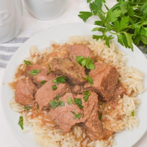 A plate of rice topped with Slow Cooker Beef Tips and Gravy, garnished with chopped parsley, with fresh parsley and salt and pepper shakers in the background.