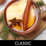 A glass of classic slow cooker cider with apple slices, a cinnamon stick, and star anise rests amidst pine branches on a white surface.