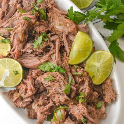 Mexican shredded beef garnished with lime wedges and parsley on a white dish, nestled next to a checkered cloth adorned with fresh parsley sprigs.