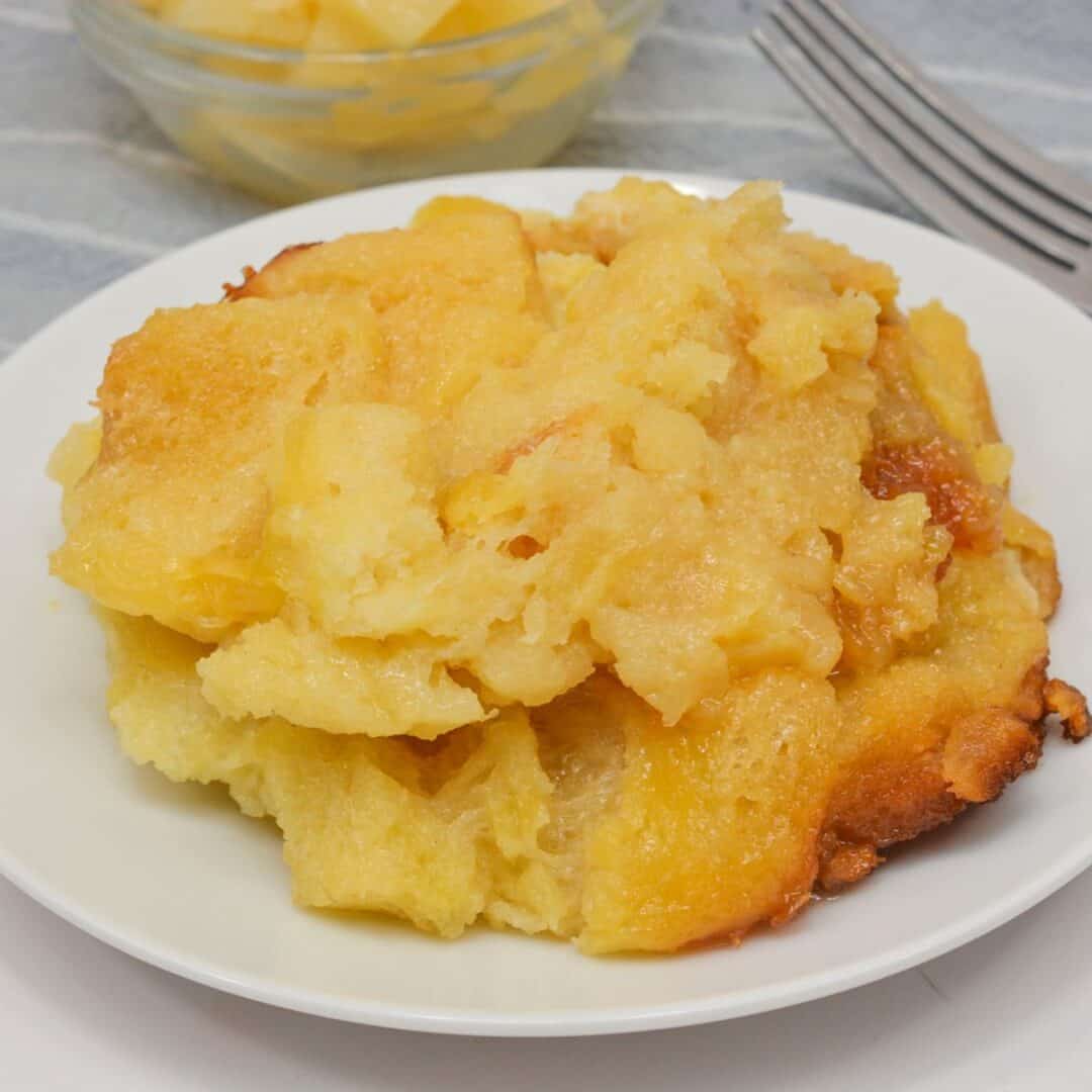 A serving of pineapple bread pudding, inspired by classic Pineapple Casserole, sits on a white plate with a bowl of pineapple chunks and two forks in the background.