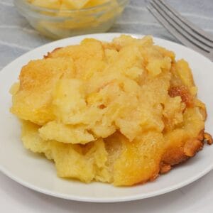 A serving of pineapple bread pudding, inspired by classic Pineapple Casserole, sits on a white plate with a bowl of pineapple chunks and two forks in the background.