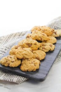 A slate serving board with a pile of soft chocolate chip cookies sits on top of newspaper-style cloth.