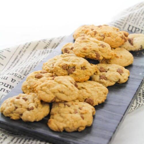 A slate serving board with a pile of soft chocolate chip cookies sits on top of newspaper-style cloth.