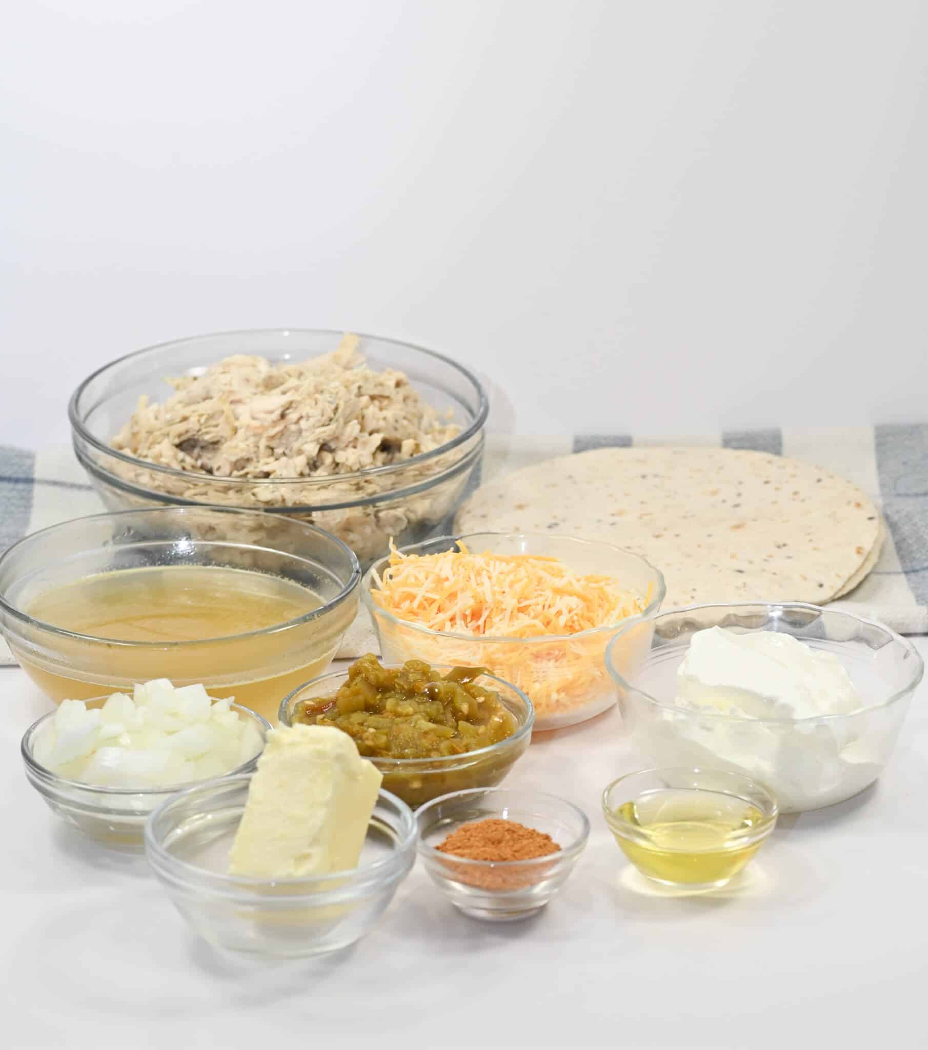 A display of ingredients features shredded chicken, tortillas, cheese, diced onions, butter, chicken broth, green chiles, sour cream, seasoning, and oil in separate bowls.
