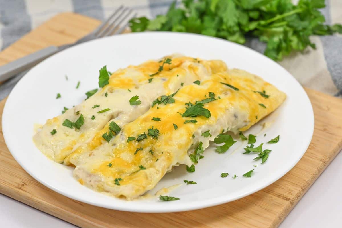 Two cheese enchiladas topped with melted cheese and garnished with cilantro on a white plate, placed on a wooden board next to a fork and a bunch of fresh cilantro, reminiscent of our delectable Sour Cream Chicken Enchiladas.