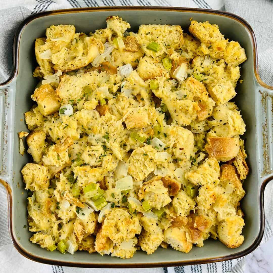 A square baking dish filled with sourdough stuffing mixed with chopped onions, celery, and herbs, set on a striped cloth.