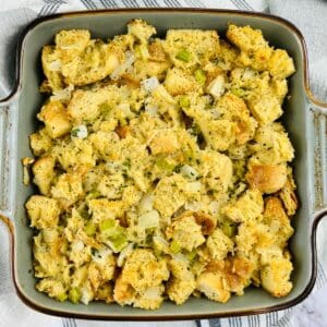 A square baking dish filled with sourdough stuffing mixed with chopped onions, celery, and herbs, set on a striped cloth.