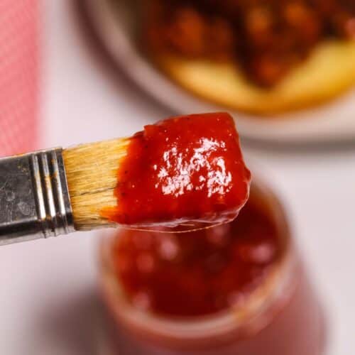 A close-up of a basting brush dipped in Chipotle BBQ Sauce, with an open jar of sauce and a sandwich bun in the background.
