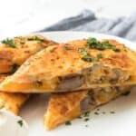 A close-up of sliced Steak Quesadillas filled with sausage and melted cheese, garnished with chopped herbs, and served with a dollop of sour cream on a white plate.