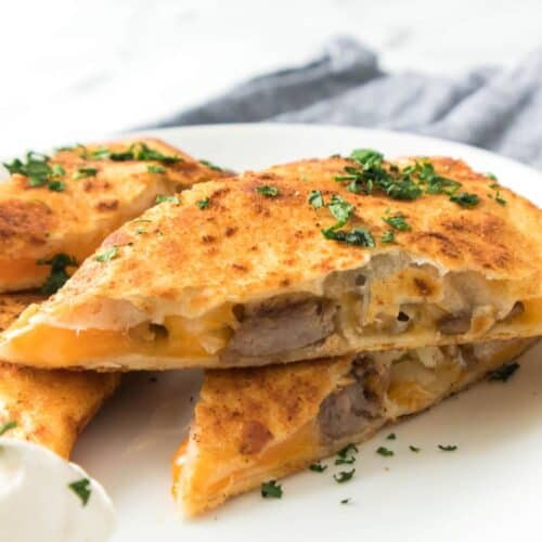 A close-up of sliced Steak Quesadillas filled with sausage and melted cheese, garnished with chopped herbs, and served with a dollop of sour cream on a white plate.