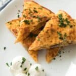 Three triangular slices are arranged on a white plate, garnished with chopped herbs, with two scoops of sour cream on the side.