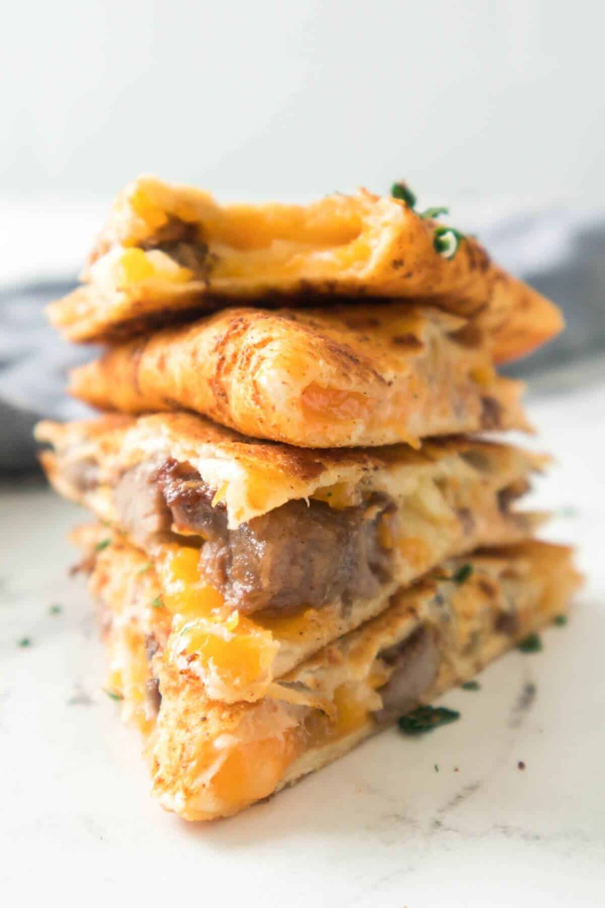 A stack of cheesy Steak Quesadillas wedges filled with tender beef, melted cheese, and fresh herbs on a light surface.
