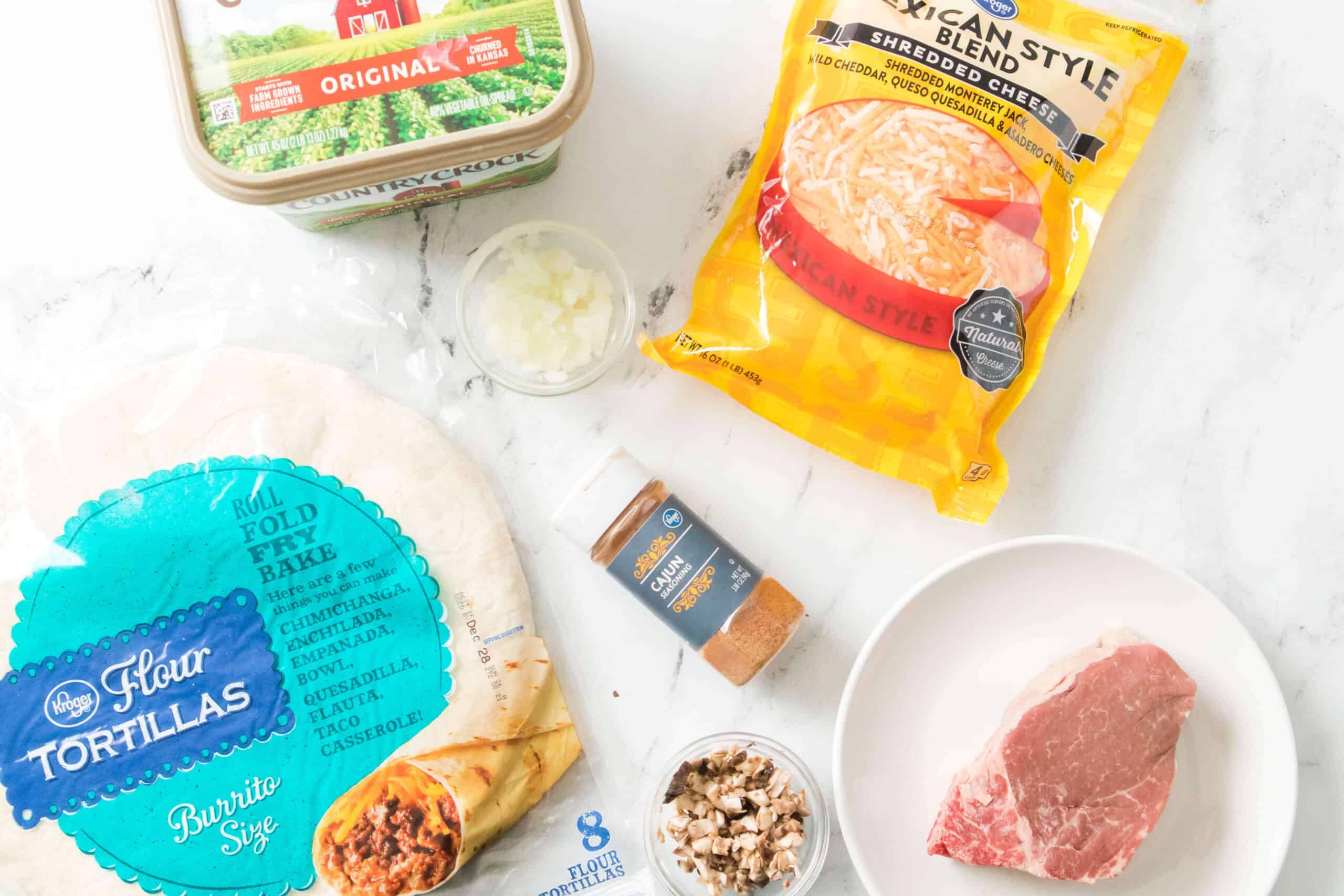 Ingredients are laid out on a white surface, featuring flour tortillas, shredded cheese, butter spread, steak, seasonings, chopped onions, and chopped mushrooms.