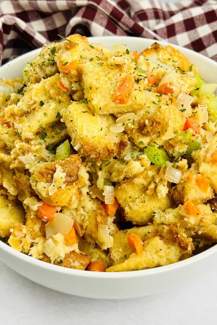A bowl filled with bread stuffing mixed with chopped celery, carrots, and onions, garnished with herbs. Inspired by a classic Stove Top Stuffing Recipe, it’s set against a checkered cloth background.
