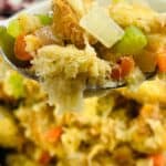 A close-up of a spoonful of bread stuffing, made using the Stove Top Stuffing Recipe, with visible chunks of bread, celery, carrots, and onions, held over a bowl of the same dish.