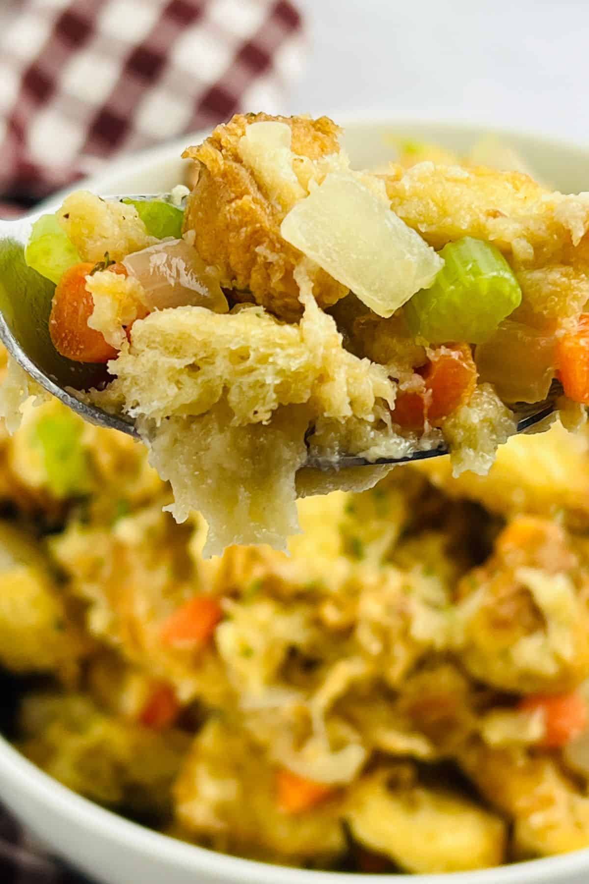 A close-up of a spoonful of bread stuffing, made using the Stove Top Stuffing Recipe, with visible chunks of bread, celery, carrots, and onions, held over a bowl of the same dish.