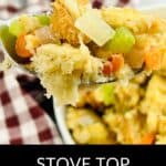 A spoonful of Stove Top Stuffing Recipe with visible bread cubes, celery, onions, and carrots is held above a bowl, with a checkered cloth in the background.