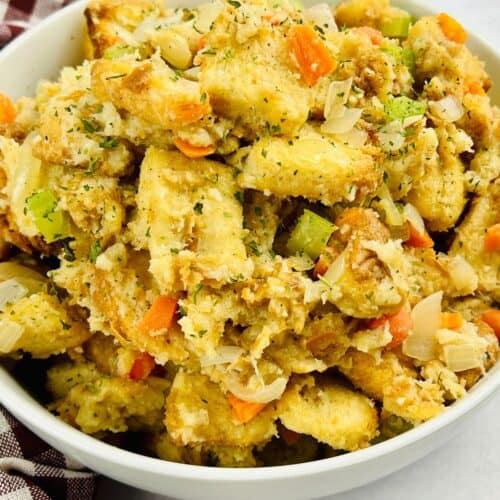 A white bowl filled with bread stuffing, made using a classic Stove Top Stuffing Recipe and mixed with chopped celery, carrots, and onions, placed beside a red and white checkered cloth.